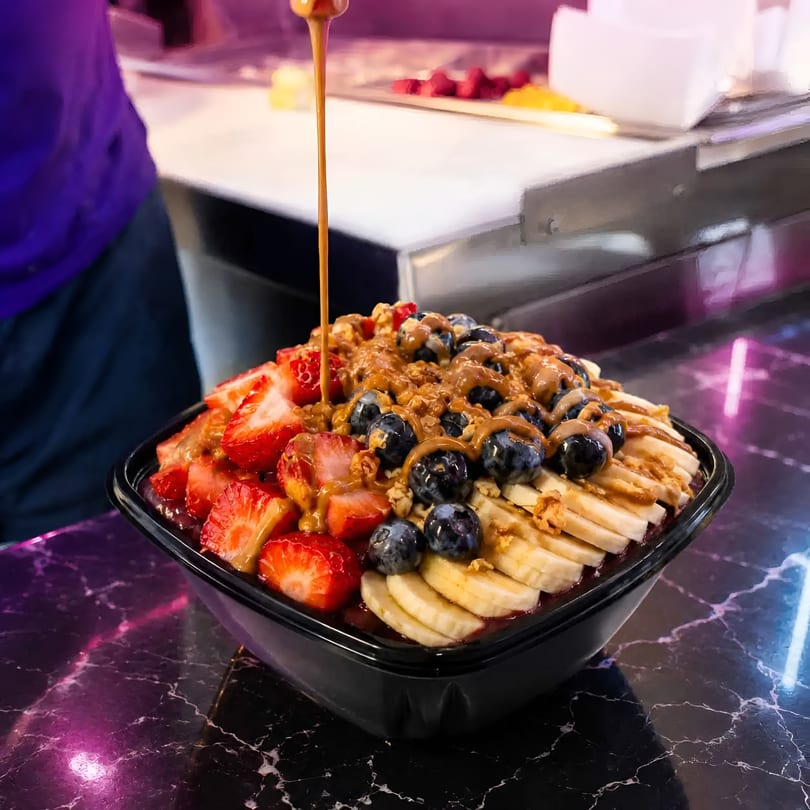 Acai bowl topped with strawberries, blueberries, bananas, granola, and a sauce drizzle on a dark counter.