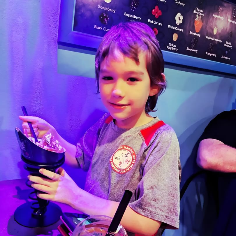 Smiling boy in purple light holds a berry dessert, with a berry menu in the background.