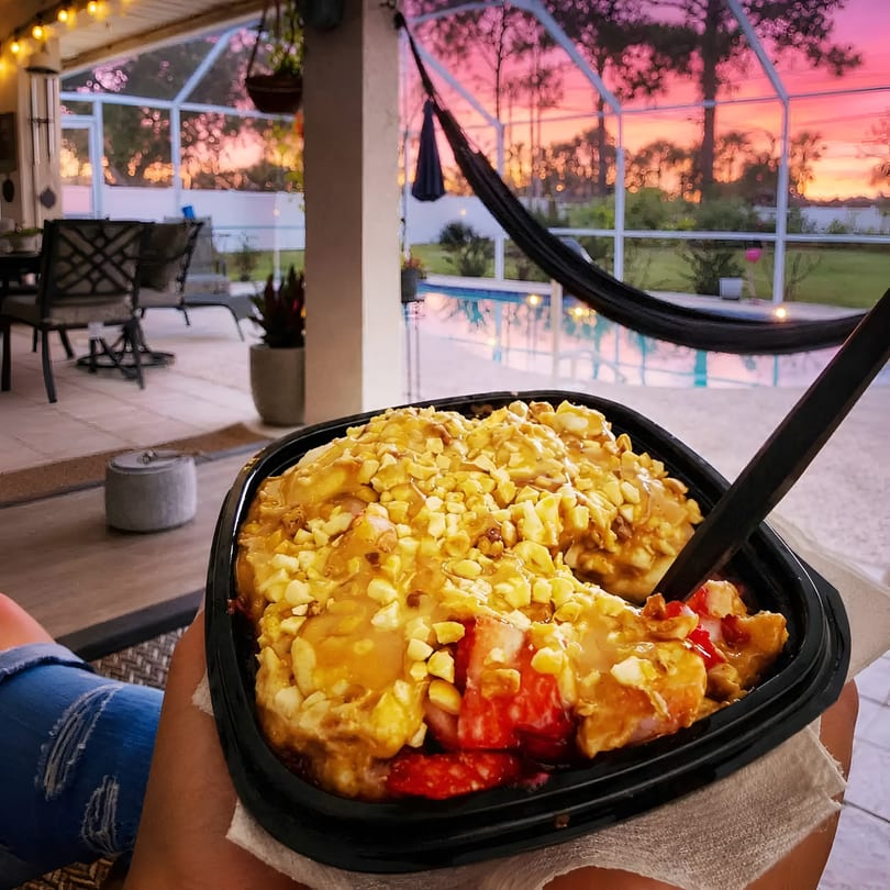 Hands holding a dessert bowl with fruit and nuts on a patio with a vibrant sunset.