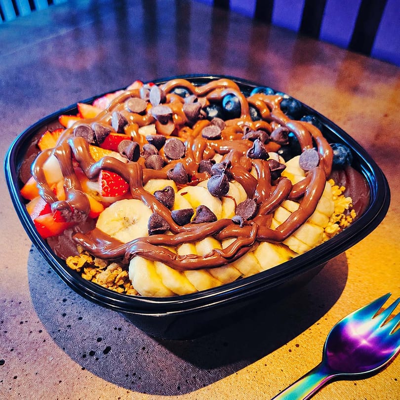 Acai bowl with bananas, strawberries, blueberries, chocolate drizzle, and chocolate chips next to a colorful spoon.