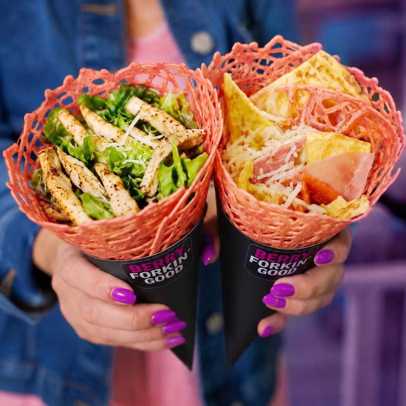 Hands hold two savory crepes in black cones, one with chicken salad, the other with ham and egg.