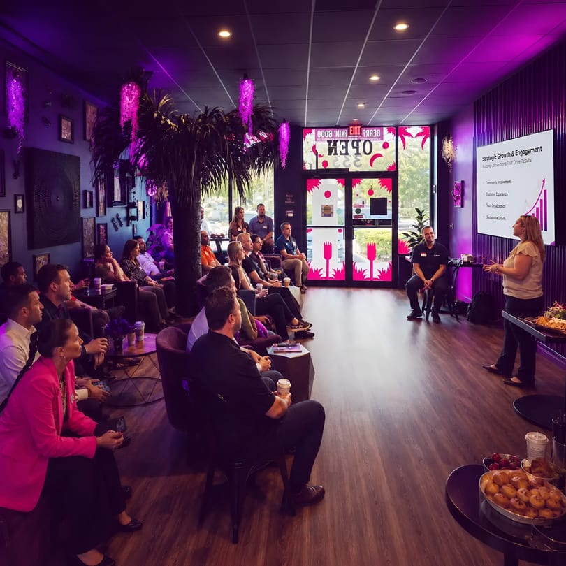 Audience watches a speaker present in a vibrant, purple-lit restaurant with snacks.