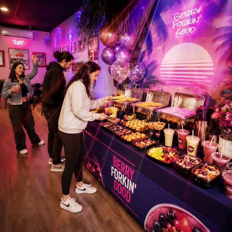 Guests enjoy a colorful fruit and smoothie buffet under neon lights at a "Berry Forkin' Good" party.