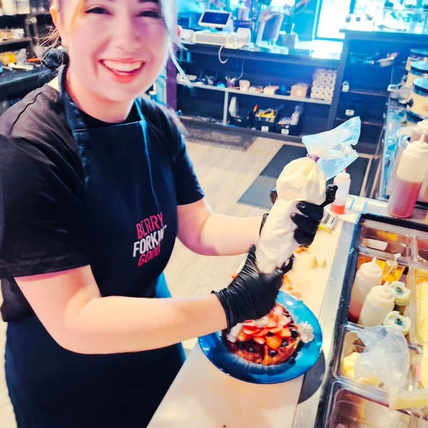 Smiling woman in a "BERRY FORK" apron piping whipped cream onto a berry waffle.