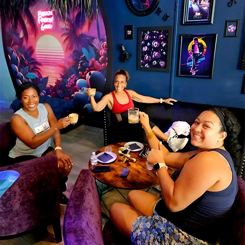 Three smiling women toast with drinks in a vibrant, neon-lit restaurant with colorful murals.