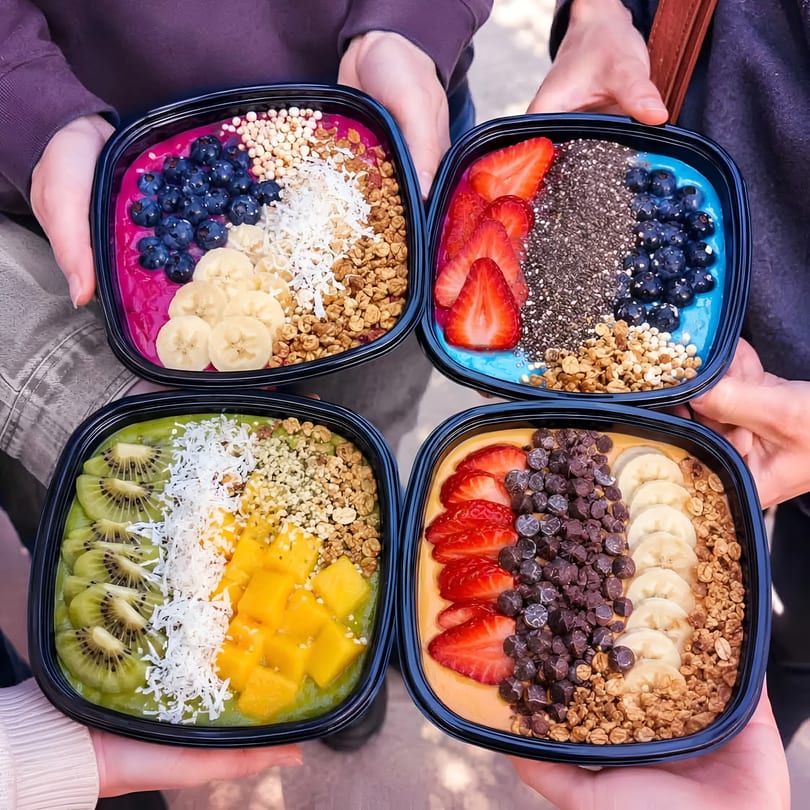 Four hands hold colorful smoothie bowls topped with various fruits, granola, and seeds.