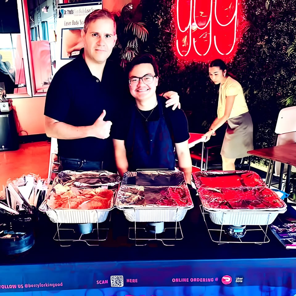 Two men pose behind a buffet table with three foil food pans, a woman works nearby.