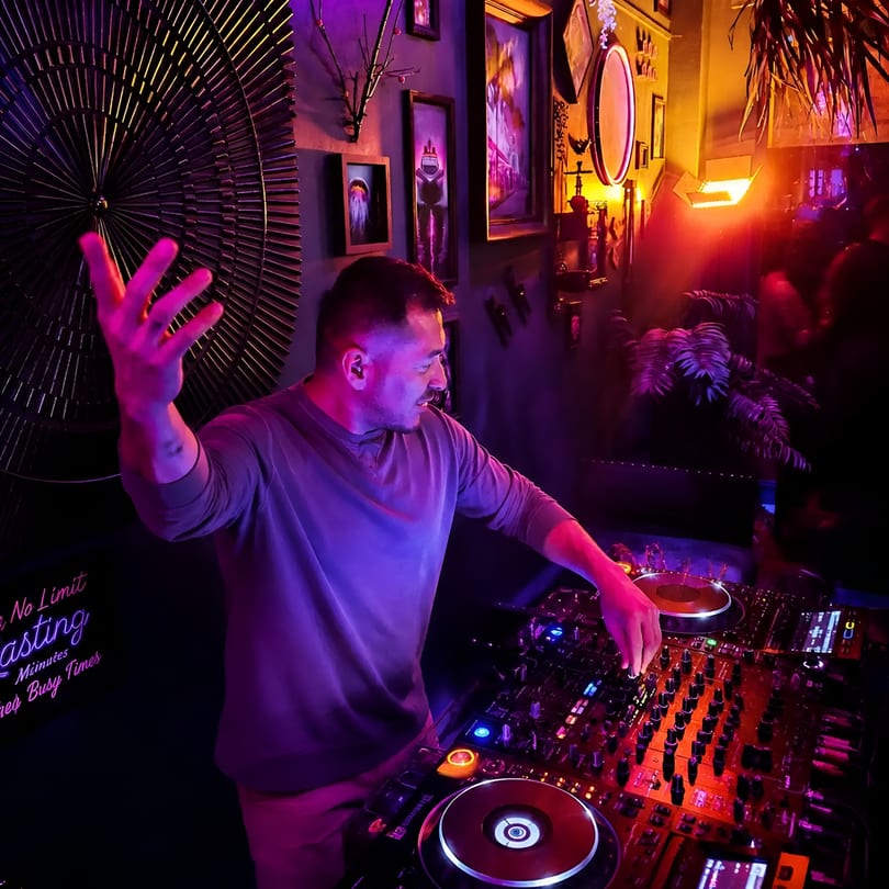 Male DJ with a raised hand mixing music on a console in a vibrant, neon-lit club.