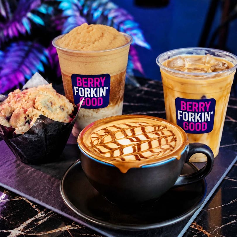 Berry Forkin' Good branded iced coffee, blended drink, caramel latte, and blueberry muffin on a dark table.