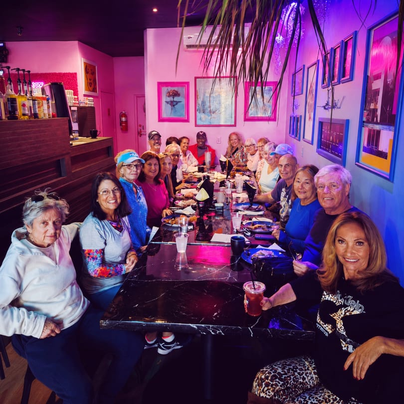 A large group of people dining at a long table in a restaurant with pink walls and blue lighting.
