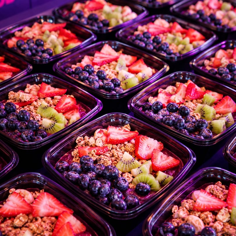 Rows of acai bowls topped with blueberries, sliced strawberries, kiwi, and granola in plastic containers.