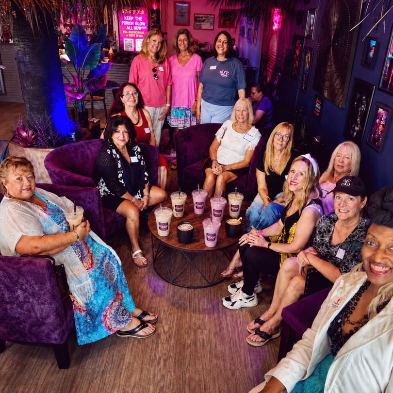 A group of women socialize in a vibrant, purple-lit lounge with drinks on a coffee table.