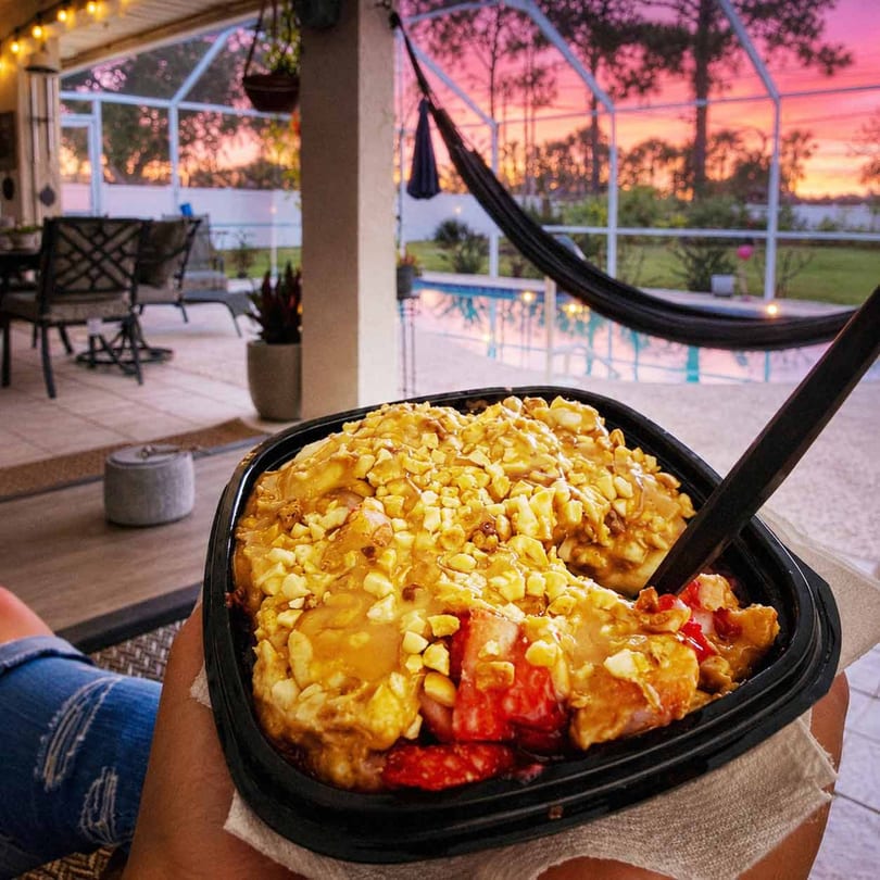 A person holds a dessert with fruit and nuts, overlooking a pool and pink sunset.