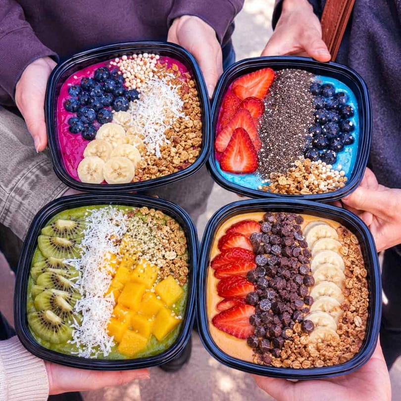 Four hands hold colorful smoothie bowls topped with various fruits, granola, and seeds.